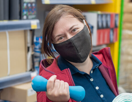 A Woman In A Medical Mask Chooses Sporting Goods In The Store. A Smiling Middle-aged Woman Holds Sports Equipment.