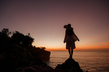 Back view young woman in straw hat standing on top of a cliff and look at the sea at sunset. Summer landscape with girl, orange sunlight