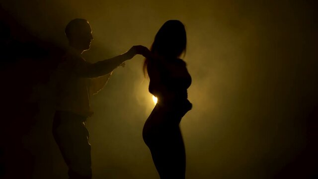 Beautiful couple dancing latin dance. Professional dancers dancing salsa in a dark smoky studio with yellow soft light. Close up shot. Silhouettes.