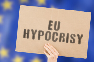 The phrase " EU hypocrisy " on a banner in men's hand with blurred European Union flag on the background. Lying. Fake. Dishonest. Untruth