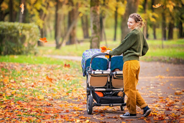Happy mom are walking in autumn park with a stroller for twins. Full-length portrait.