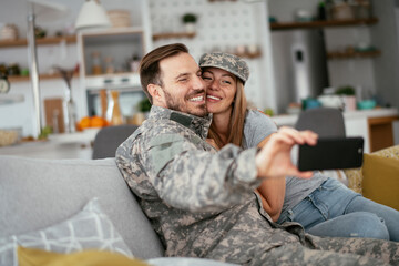 Young soldier taking selfie with his wife at home. Loving couple sitting on sofa in living room..