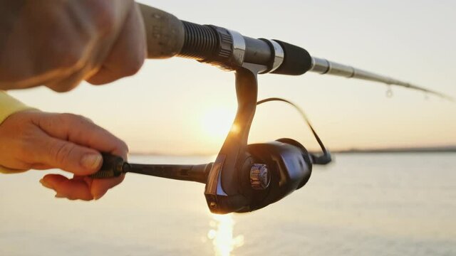 Handle rotation with reel of fishing rod against of orange sunset slow motion. Woman hobby fishing on sea tightens a fishing line reel of fish summer. Lens flare. Calm surface sea. Bright disk of sun