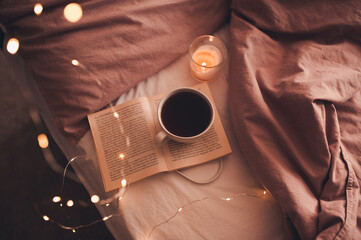 Cup of fresh hot coffee staying on open paper book with burning candle and glowing Christmas lights...