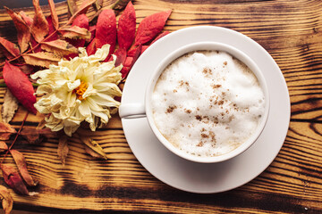 Coffee cup on the autumn fall leaves and dried flower and wooden surface background.  White cup of coffee latte with cinnamon. Seasonal fall concept. Top view, flat lay.