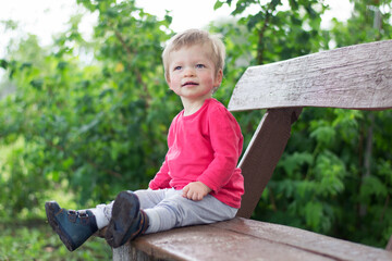 A small, smiling child sits on a bench in a summer Park. There is a green shrub in the background.