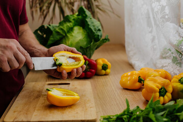 Happy handsome man is cooking in the kitchen by the window. Cut vegetables. Fresh bell peppers,...