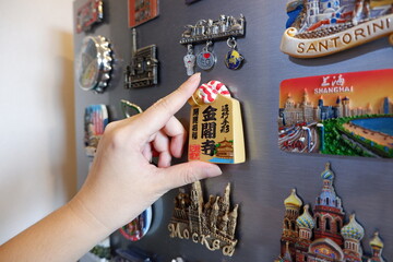 Putting a new magnet on fridge door, closeup hand. Different travel souvenir magnet on the fridge - obrazy, fototapety, plakaty