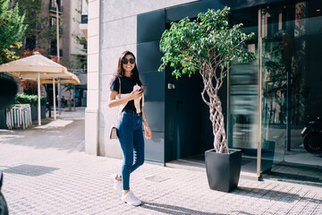 Portrait of cheerful asian female walking on street during sunny day in summer using mobile phone, smiling beautiful woman 20s millennial strolling with smartphone connected to 4G for navigation