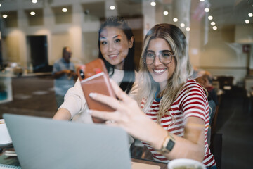 Positive caucasian female freelancer making picture with asian female colleague cooperate on freelance online project, smiling hipster girl students taking photo while learning together using laptop