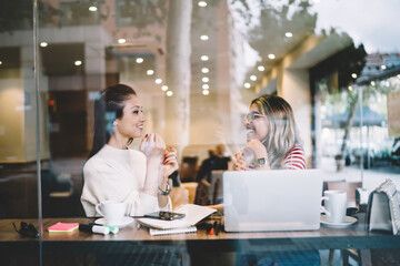 Beautiful positive female colleagues having conversation about remote work in cafeteria using laptop computer, smiling hipster girl students sitting near window in cafe together communicating