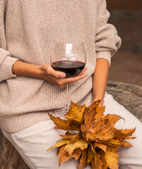 A women's hands holding a glass of red wine and an autumn bouquet of yellow fall leaves. Beige trendy total look with warm sweater and trousers. Trendy autumn concept in pastel colors. Autumn mood.