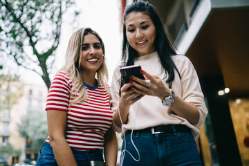 Half length portrait of Spanish millennial girl smiling at camera while Asian best friend choosing audio set for listening during together pastime, happy tourists tracking online guide on cellphone