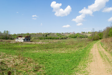 There is dry grass in the foreground. On the middle plan on the green field is an old, destroyed house. In the background is a hill with rural houses.