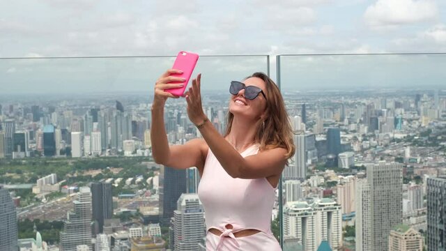 Young happy smiling caucasian woman traveller in fitting dress and sunglasses on high floor in bangkok, holding phone   