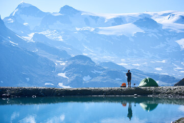 Male tourist enjoying scenery, standing and talking by radio near tent in empty rocky alpine mountains near lake with fresh clear water. Concept of beauty nature, camping and travelling in Alps
