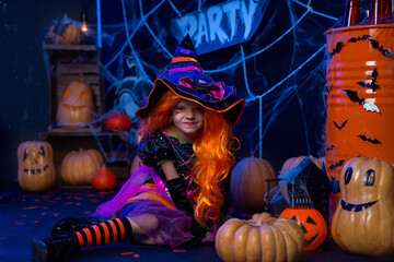 Little beautiful girl in a witch costume celebrates Happy Halloween party in interior with pumpkins.