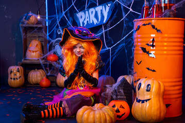 Little beautiful girl in a witch costume celebrates Happy Halloween party in interior with pumpkins.