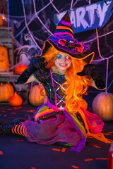 Little beautiful girl in a witch costume celebrates Happy Halloween party in interior with pumpkins.