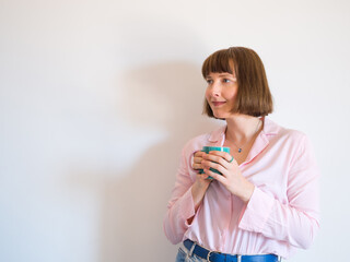 Young woman wearing jeans and pink shirt drinking coffee and smiling over white wall background.
