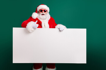 Photo of retired stylish grandfather grey hair beard hold white board thumb-up assure chosen object wear red santa x-mas costume coat gloves sunglass headwear isolated green color background © deagreez