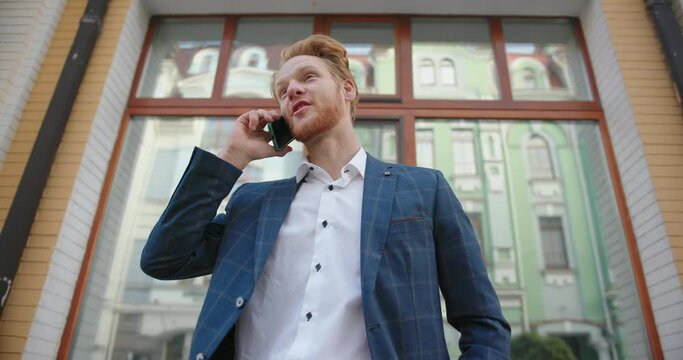 Red haired man in jacket answers call and talks on cellphone by large window with building reflection on street low angle shot