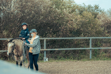 Happy kid boy riding a horse training with instructor. The boy learns to ride a horse. Hippotherapy for the development of the baby. A child in a warm suit and helmet for safety. Safety quarantine