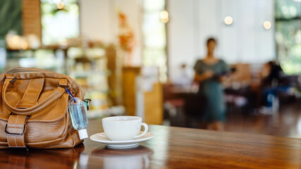 Mini alcohol gel bottle to kill Corona Virus(Covid-19) hang on a brown leather shoulder bag with hot coffee on table in cafe.New normal lifestyle.Health care concept.Selective focus on alcohol gel