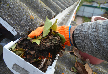 A man is cleaning a clogged roof gutter from dirt, debris and fallen leaves to prevent water damage...