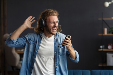 Handsome long haired curly man with headphones listening music,singing and dancing.Funny emotional smiling man with earphones and mobile phone relax at home.Enjoy listening to music.Stress management.