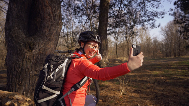 Young caucasian woman athlete tourist cyclist uses hand smart phone photo of herself selfie sitting near tree in coniferous forest outside the city. Sportswoman taking selfie with her mountain bike