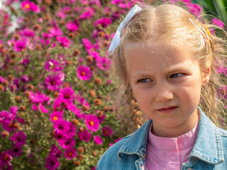 Beautiful sad blond little girl smelling flower and feeling upset. Kids pollen allergy. Sick child grimaces and sneezes on blurred nature background. Allergic rhinitis, plants blooming.