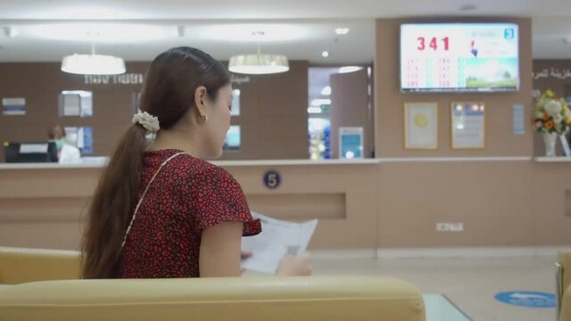 asian woman sitting in hospital  waiting lounge. young adult female waiting for queue number call up from monitor at payment counter. she is going to claim insurance. 