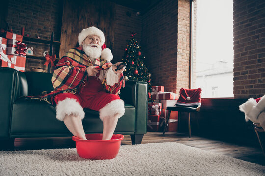 Full Length Photo Funny Grey White Beard Hair Santa Claus Sit Couch Rest X-mas Delivery Present Watch Tv Switch Remote Control Soak Hot Water Feet Wear Headwear In House Indoors Christmas Tree