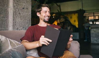 Cheerful emotional caucasian male laughing during reading stories enjoying recreation and hobby, positive hipster guy in casual wear holding black menu choosing food in cafeteria enjoying free time