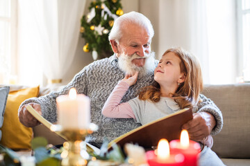 Small girl with senior grandfather indoors at home at Christmas, looking at photographs.