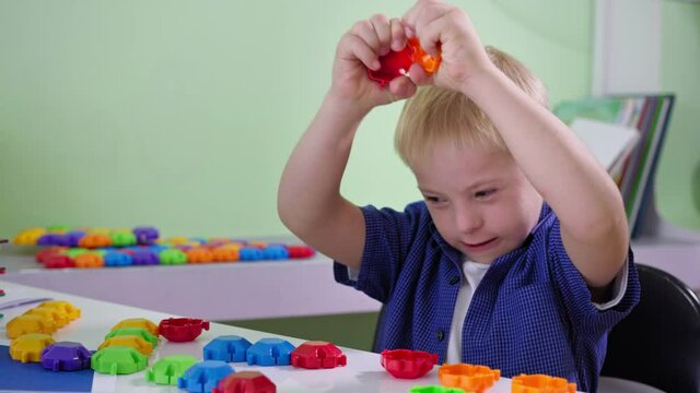 interested child with Down syndrome disassembles and plays with a colorful constructor sitting at a table at home close up