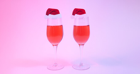 Christmas and new year holiday greeting card concept with two wine glasses with rose wine decorated with Santa hats on multicolored background in neon lights w. Copy space.