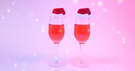 Christmas and new year holiday greeting card concept with two wine glasses with rose wine  decorated with Santa hats on pink background in neon lights with multicolored snowflake bokeh. Copy space.