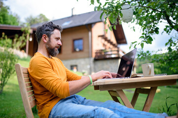 Side view of man with laptop working outdoors in garden, home office concept.