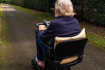 An old lady in a blue jacket riding a mobility scooter on a quiet lane from behind.