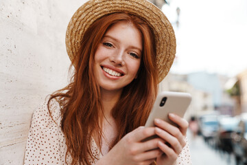Image of smiling ginger girl using cellphone while walking on city