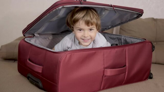 Little Kid Is Playing Inside Suitcase. Little Kid in Travel Buggage. Packed for Vacation. Happy Child Boy Tourist Looks Out of Red Suitcase For Travel Vacation Adventure. Kid Having Fun at Home.
