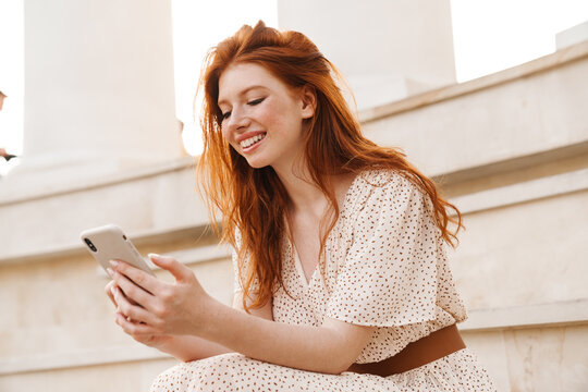 Image of cheerful ginger girl using mobile phone while sitting on stairs