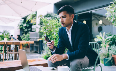 Serious hindu male entrepreneur in formal trendy outfit sitting outdoors on terrace working remotely on laptop computer, pensive businessman checking banking transaction notification on smartphone