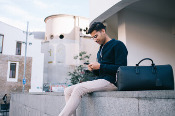 Handsome young confident male in trendy outfit sitting outdoors on uran setting using mobile phone for checking location during trip,hindu hipster guy in stylish wear with luggage dialling number