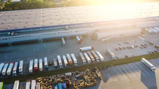 Logistics park with a warehouse - loading hub. Semi-trucks with freight trailers standing at the ramps for loading/unloading goods at sunset. Aerial hyper lapse - motion time lapse
