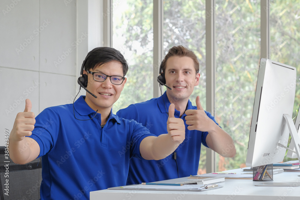 Wall mural Smiley face asian male operator giving thumbs up and looking to camera with the colleague at the office space background.