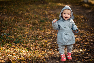 Happy child on the street. Baby smiles in the autumn Park. A kid in a gray coat and pink boots. Brown leaves on the ground. Warm autumn.