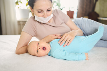 Obraz na płótnie Canvas Beautiful mother in protective face mask holding her little cute newborn son protecting him from viruses and infections. Coronavirus covid-19 prevention. Home quarantine. Happy family at home.
